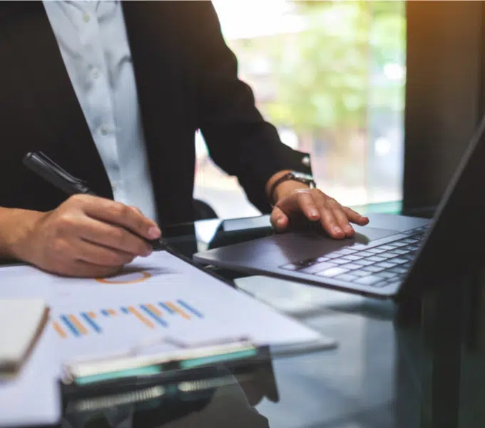 Person in business attire uses a laptop and reviews printed financial data at a desk.